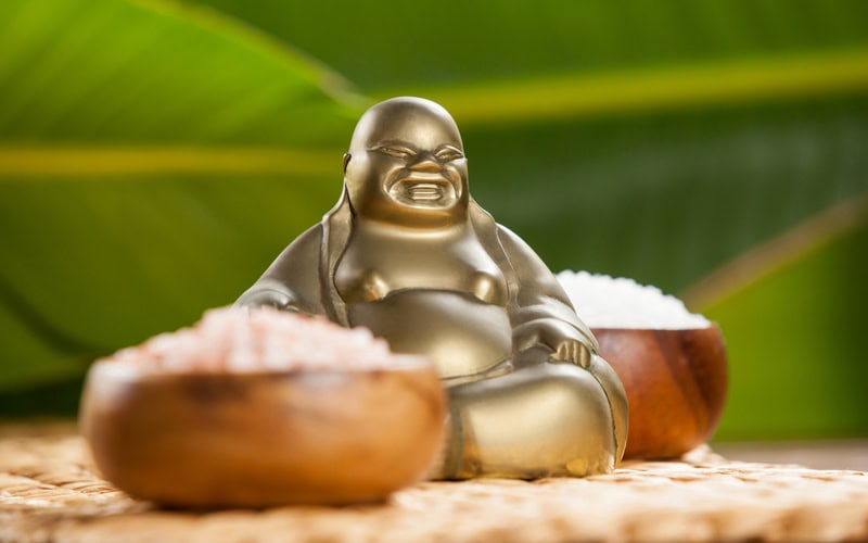 Laughing buddha figurine and sea salt in wooden bowl on mat